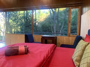 A room with a large bed, a small table, and a view of a forest through large windows. - Pine Trails Jungle Camp (Kasauli)