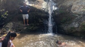 Un ambiente naturale con una cascata, terreno roccioso e persone che interagiscono con l'acqua.