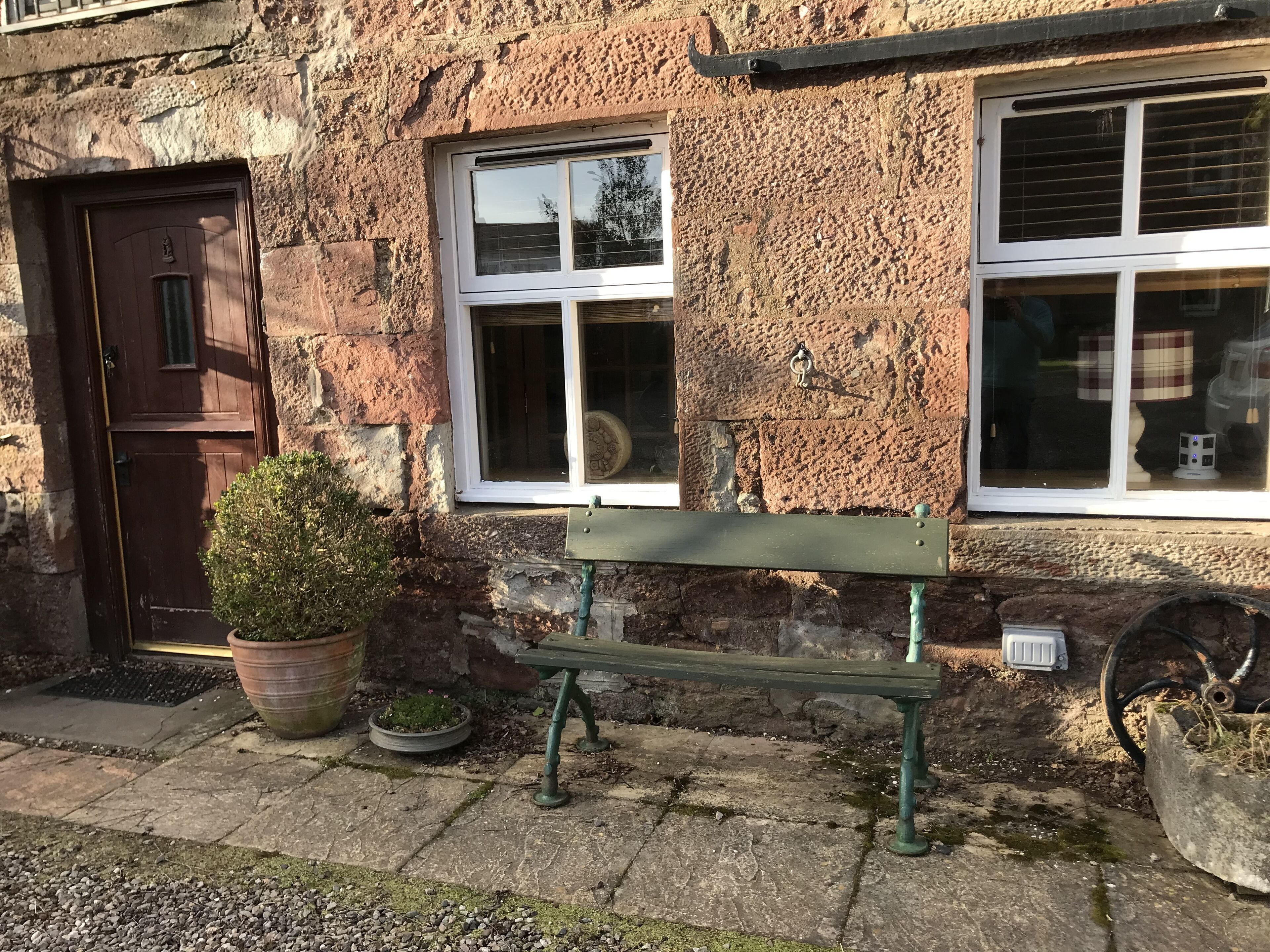 A rustic stone building with a wooden door, two windows with shutters, a bench, and potted plants.