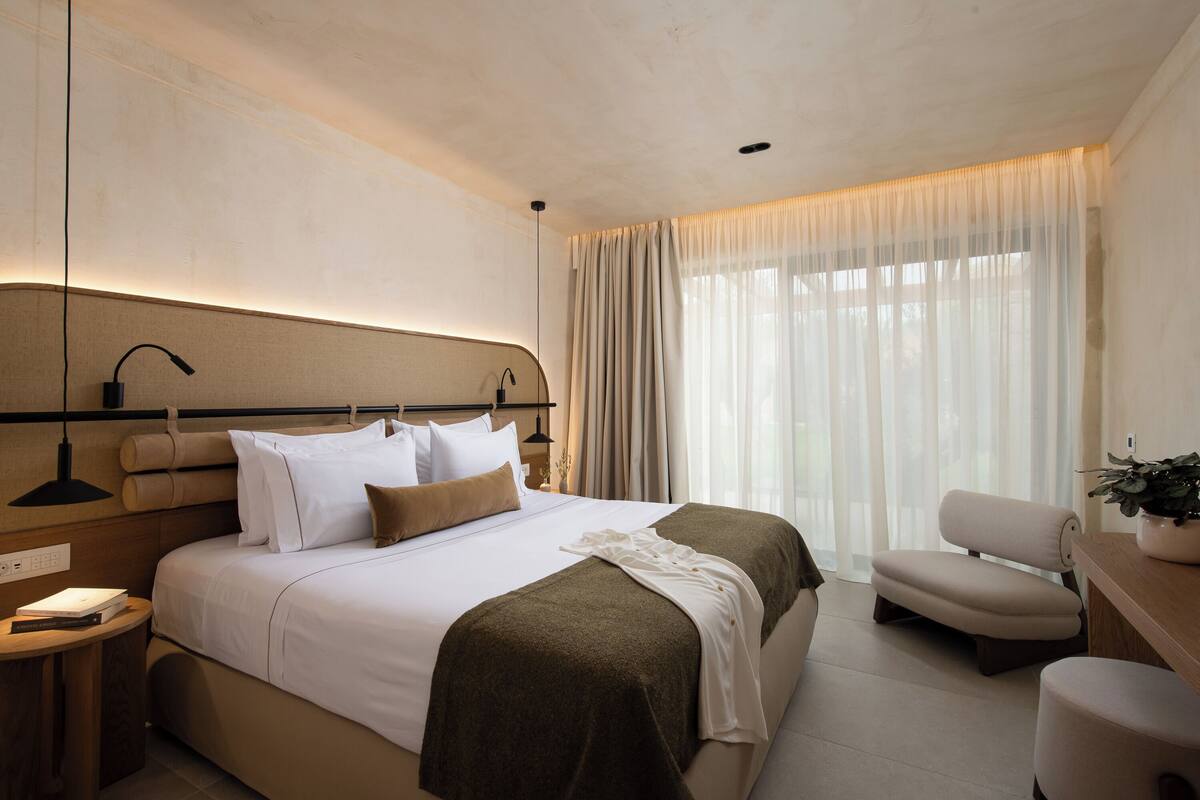 A modern hotel room with a large bed, a bedside table, a chair, and a desk.
