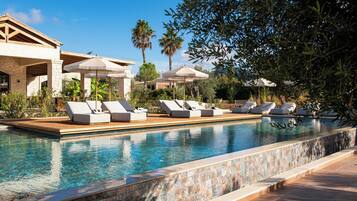 Piscina con sillas de descanso y sombrillas, rodeada de palmeras y un edificio de piedra.