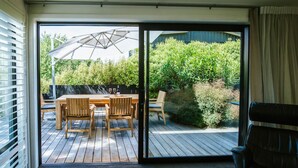 Een open glazen schuifdeur die leidt naar een terras met een houten tafel en stoelen, een pergola en uitzicht op een tuin en bomen.