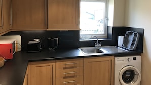 Una cocina compacta con armarios de madera, lavadora integrada, ventana con vistas y encimera negra.