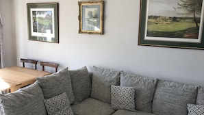 Una sala de estar con un sofá gris, una mesa de comedor de madera y tres cuadros con paisajes en la pared.