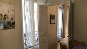 Habitación de hotel con piso de madera, una cama, una mesa y una silla. Se ve una ventana con cortinas y un radiador debajo.