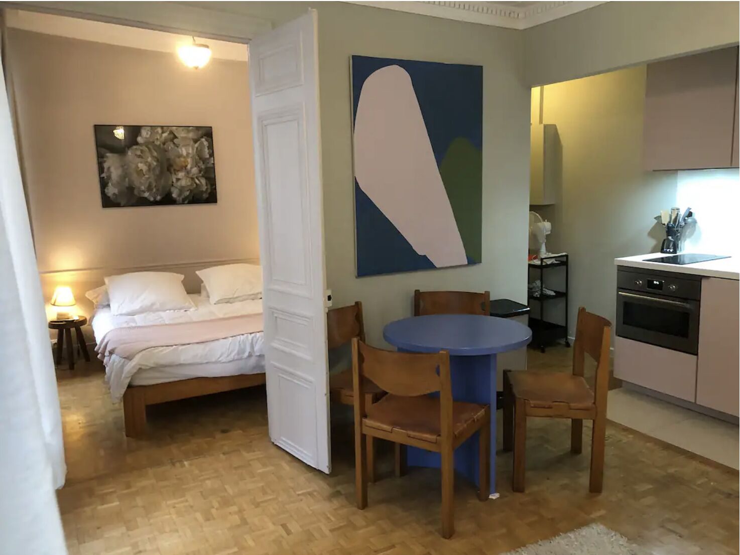 Een slaapkamer met een bed, een nachtkastje met een lamp, een groot abstract schilderij, en een deur die leidt naar een eethoek met een blauwe tafel en houten stoelen.