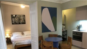 Un dormitorio con cama, una mesita de noche con lámpara, un cuadro abstracto grande y una puerta que da a un comedor con mesa azul y sillas de madera.