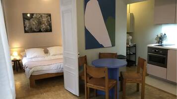 Een slaapkamer met een bed, een nachtkastje met een lamp, een groot abstract schilderij, en een deur die leidt naar een eethoek met een blauwe tafel en houten stoelen.