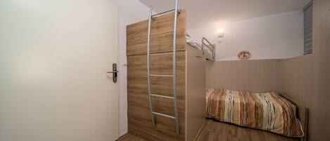 Una habitación con un armario de madera, una escalera, una cama con colcha a rayas y un estante fijado a la pared.