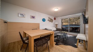 Ein gemütliches Zimmer mit einem Holztisch, Stühlen, einer Couch und einem kleinen runden Tisch. Ein Fenster bietet Blick auf Bäume und eine Terrasse.