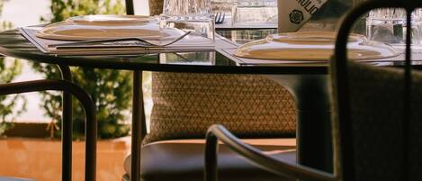 A dining table set outdoors with metal chairs, a cushioned bench, and a view of the sea.