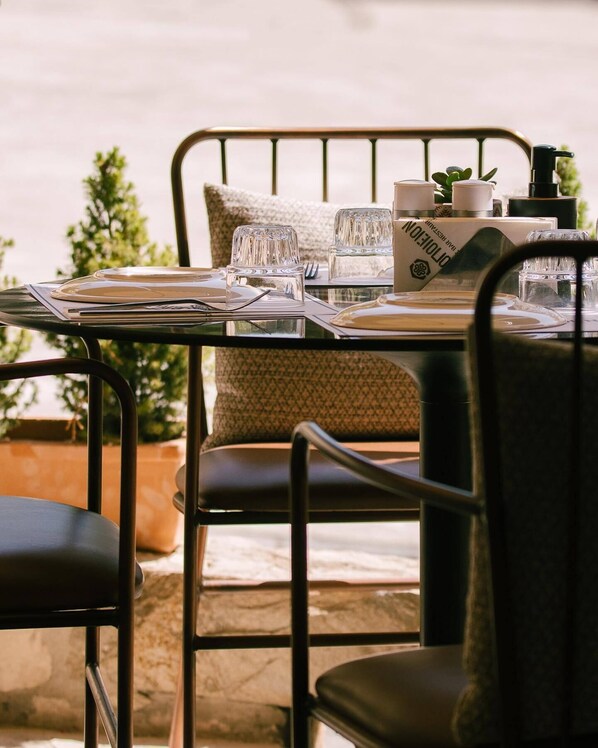 Une table à manger installée à l’extérieur, avec des chaises en métal, un banc rembourré et une vue sur la mer.