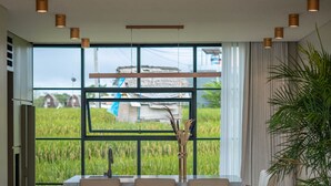 A modern dining area with a large window, a table with chairs, and a potted plant. - Vesna Townhouse 7  (Canggu)