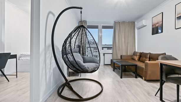 A modern living room with a hanging chair, a sofa, a dining table, and a view of the city through a large window. - RentPlanet - Apartament Muranów (Warsaw)