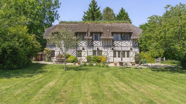 Une maison traditionnelle à colombages, dotée d’un toit de chaume, entourée d’une pelouse bien entretenue et d’arbres.