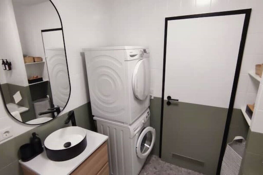 A compact bathroom with a white sink, a mirror, a washing machine, and a door.