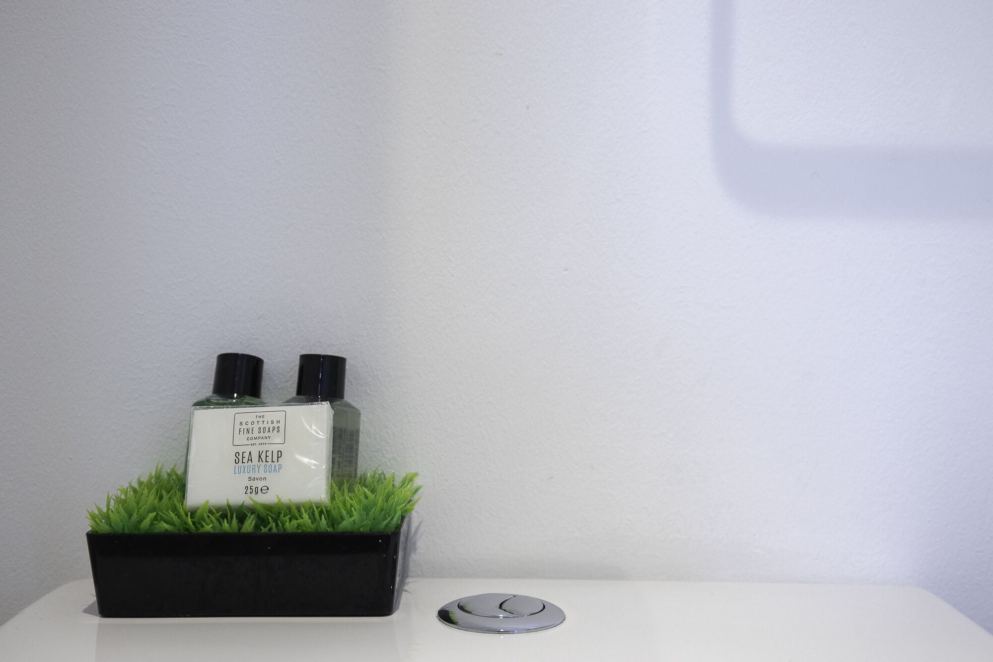 Dos jabones Sea Kelp Luxury de The Scottish Fine Soaps Company en un macetero negro con hierba verde, sobre una superficie blanca junto a un objeto redondo.