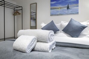A neatly made bed with grey bedding, a folded white towel, and a painting of a sailboat on the wall. - Upper Rock Brighton Apartments (Brighton)