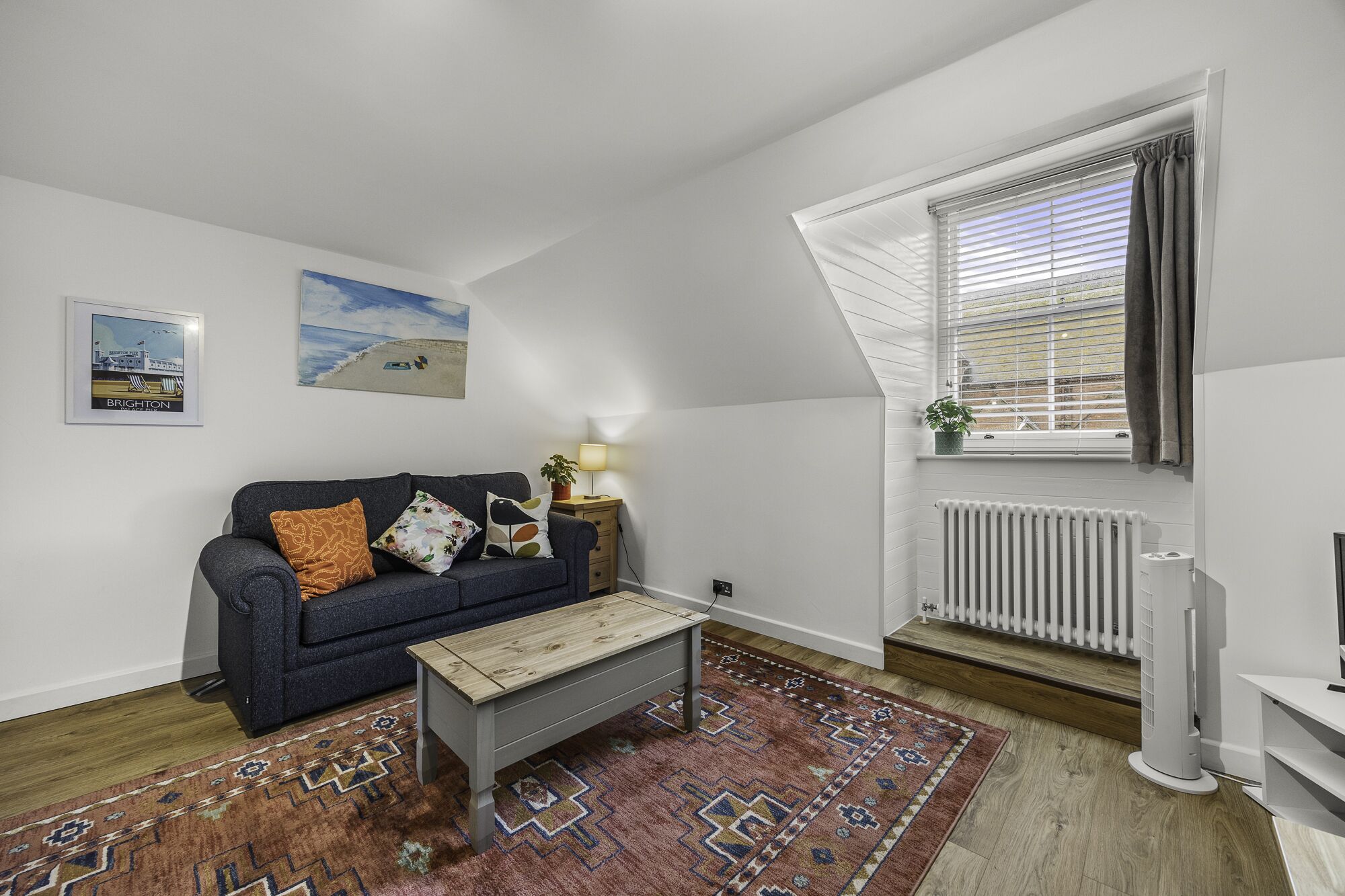 Ein Wohnzimmer mit einem dunkelblauen Sofa, einem Holztisch, einem Teppich mit Muster, einer Heizung und einem Fenster mit Jalousien.