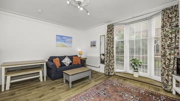 Un salon comprenant un canapé, une table basse, un tapis à motifs et une grande fenêtre avec des stores.