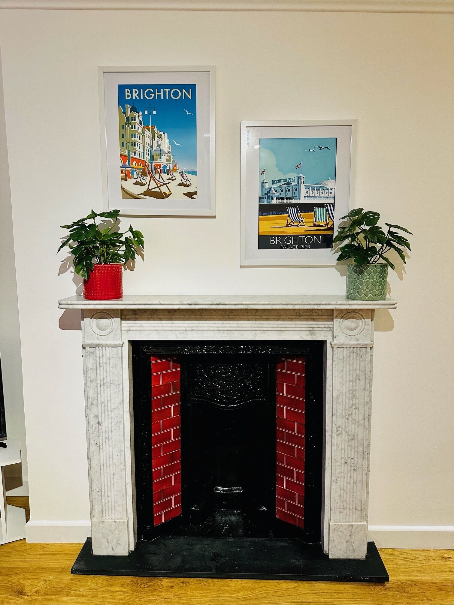 A fireplace with a white mantel and red brick interior, flanked by two potted plants. Above the mantel, two framed posters depict Brighton landmarks.