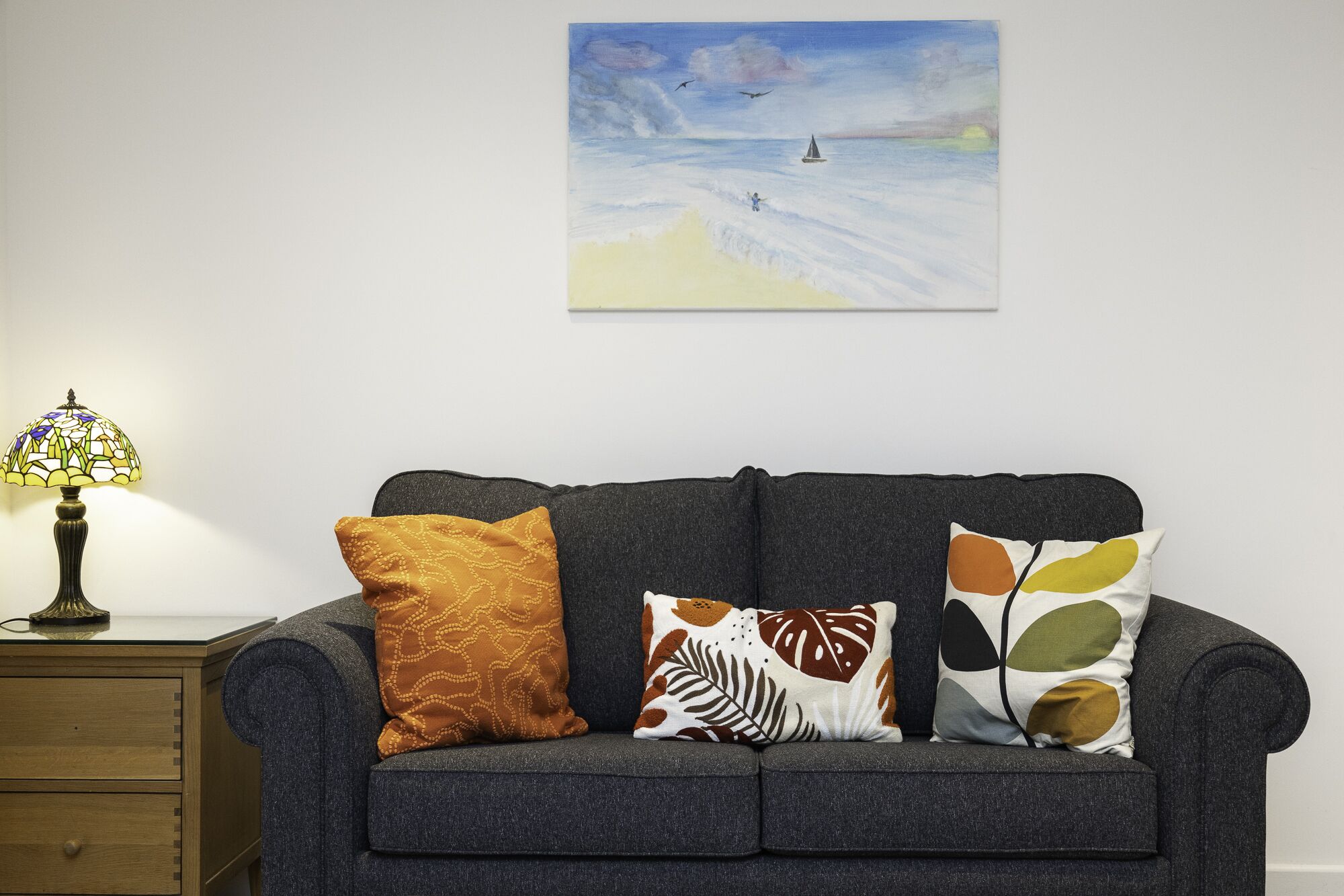 A dark grey sofa with three patterned cushions, a small wooden nightstand with a lamp, and a painting of a seascape above the sofa.
