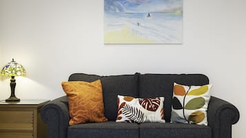 A dark grey sofa with three patterned cushions, a small wooden nightstand with a lamp, and a painting of a seascape above the sofa.