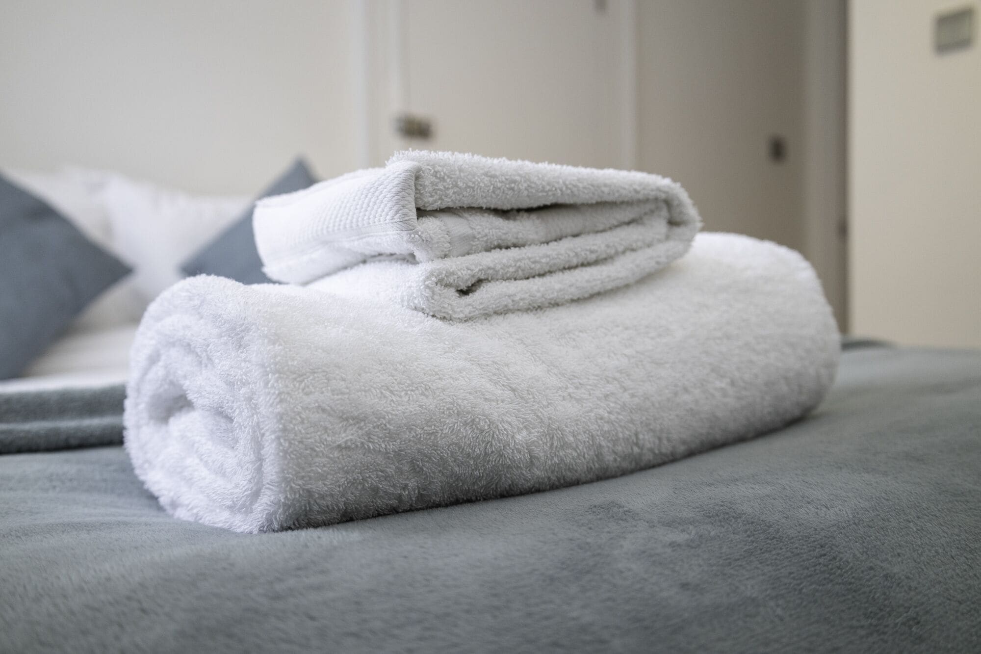 Una toalla blanca doblada con cuidado sobre una cama gris en una habitación de hotel.