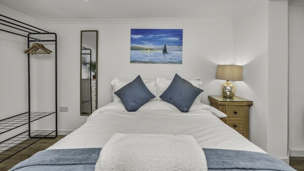Une chambre bien rangée, avec un lit, une table de chevet dotée d’une lampe, un portant à vêtements, un miroir et un tableau accroché au mur.