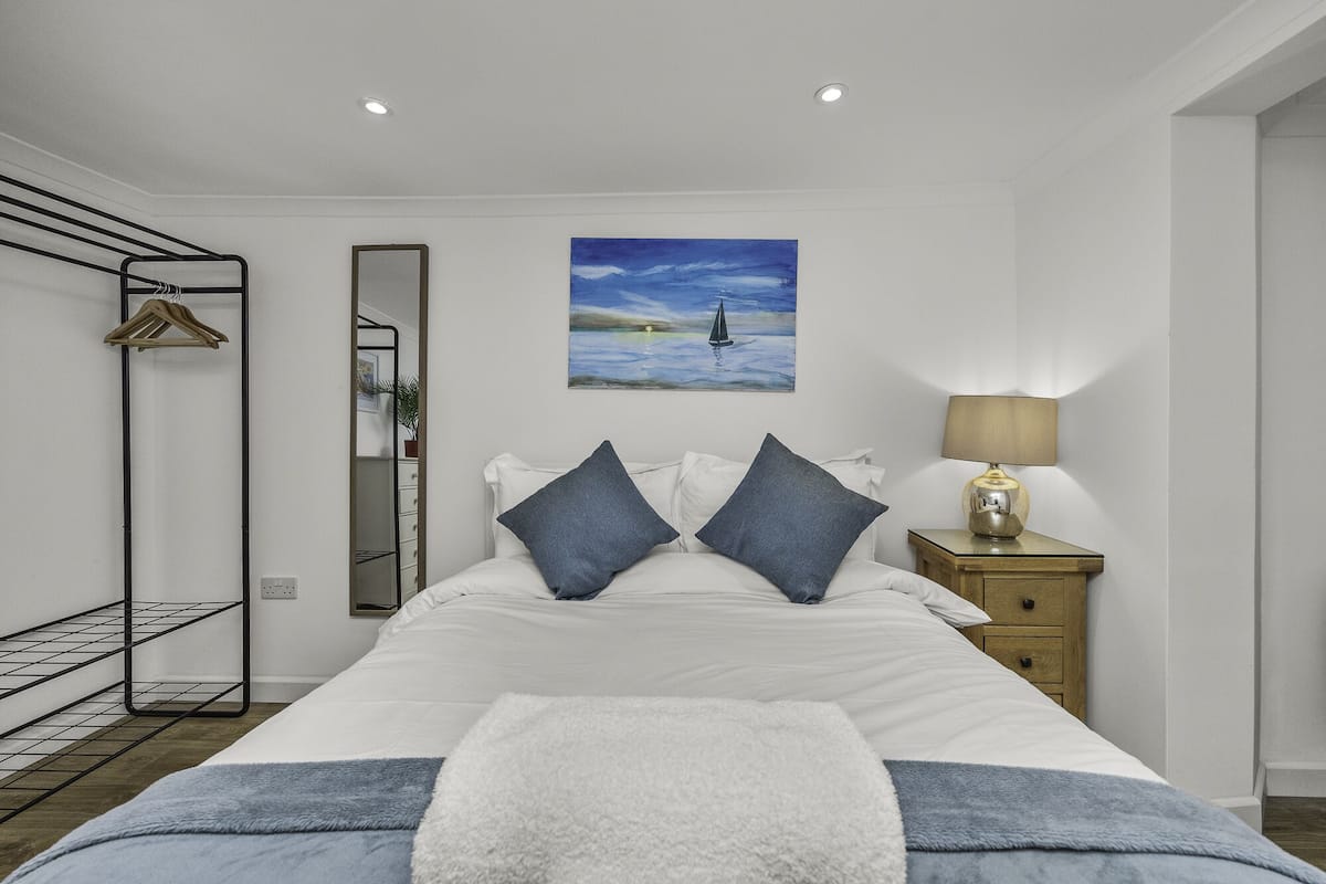 Un dormitorio ordenado con una cama, una mesita de noche con lámpara, un perchero, un espejo y un cuadro en la pared.