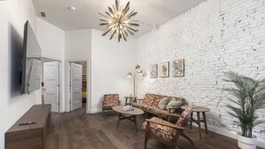 Un salon moderne avec un mur en briques blanches, un ensemble de canapés, une table basse, un téléviseur et une plante.