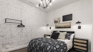Une chambre avec un mur en briques blanches, un lit recouvert d’une couvre-lit à motifs floraux noir et blanc, une tête de lit noire, une table de chevet avec une lampe et des œuvres d’art accrochées au mur.