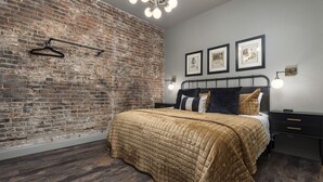 A bedroom with a brick wall, a bed with a quilted cover, a nightstand, and a wall-mounted light fixture. - Penthouse Grand Executive Suite (Louisville)