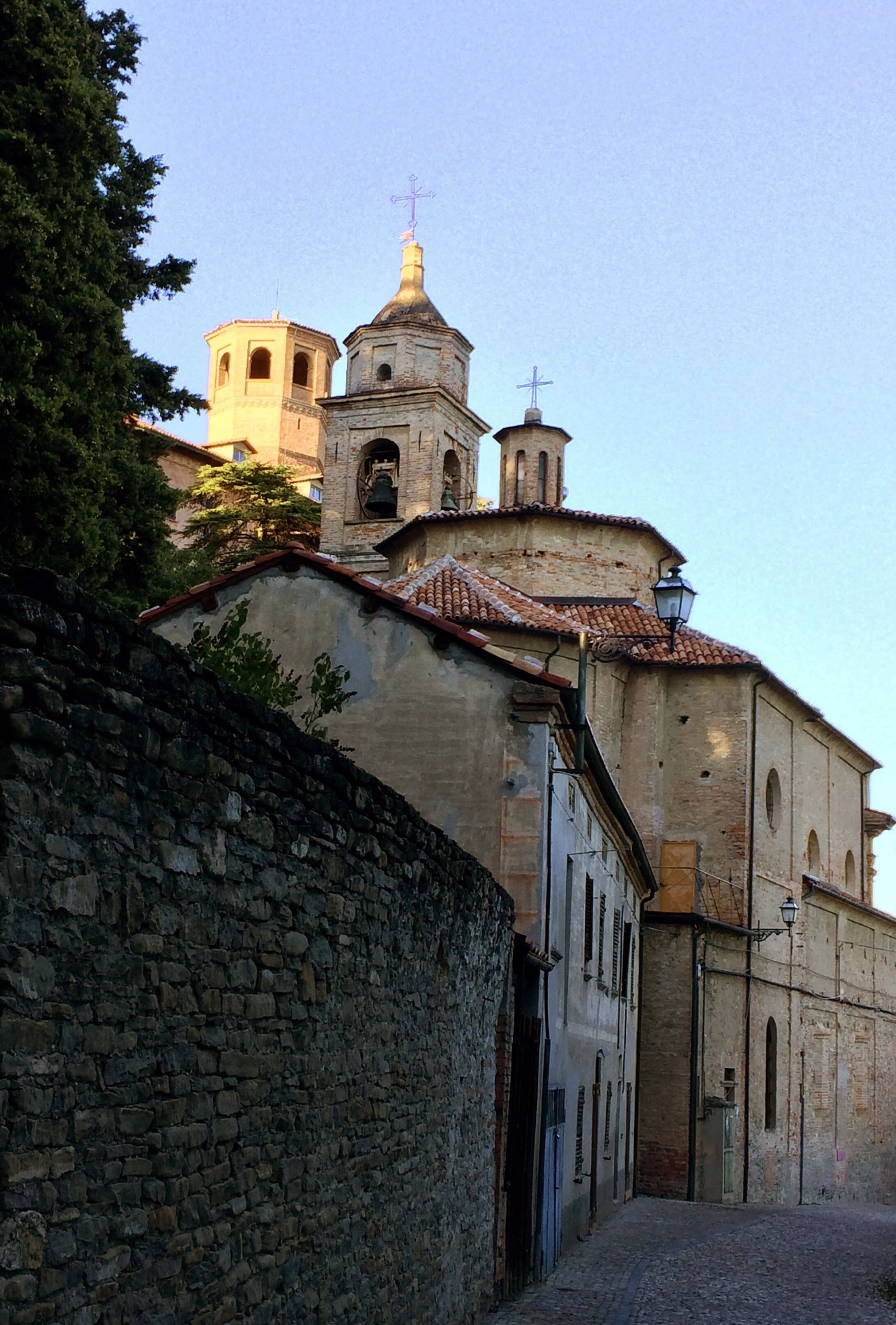 A narrow cobblestone street flanked by historic buildings with a bell tower and a cross on top.