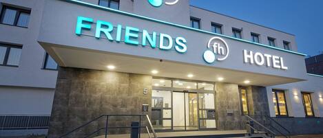 Un hotel moderno con un llamativo letrero que dice 'FRIENDS HOTEL', un logotipo con 'fh' y un diseño circular, y una escalera que conduce a la entrada.