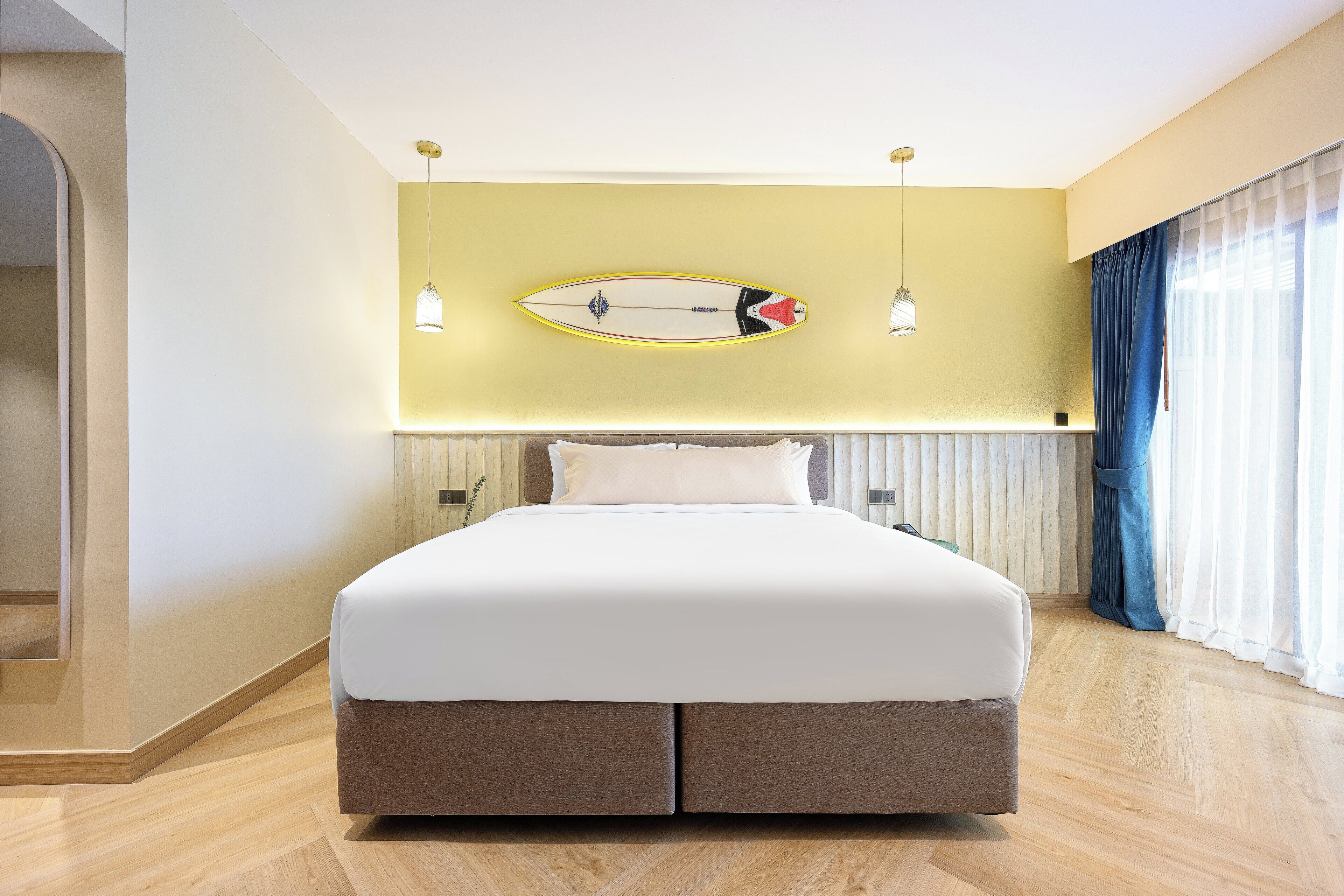 Une chambre d’hôtel moderne avec un grand lit, une planche de surf accrochée au mur et une vue sur l’océan par la fenêtre.