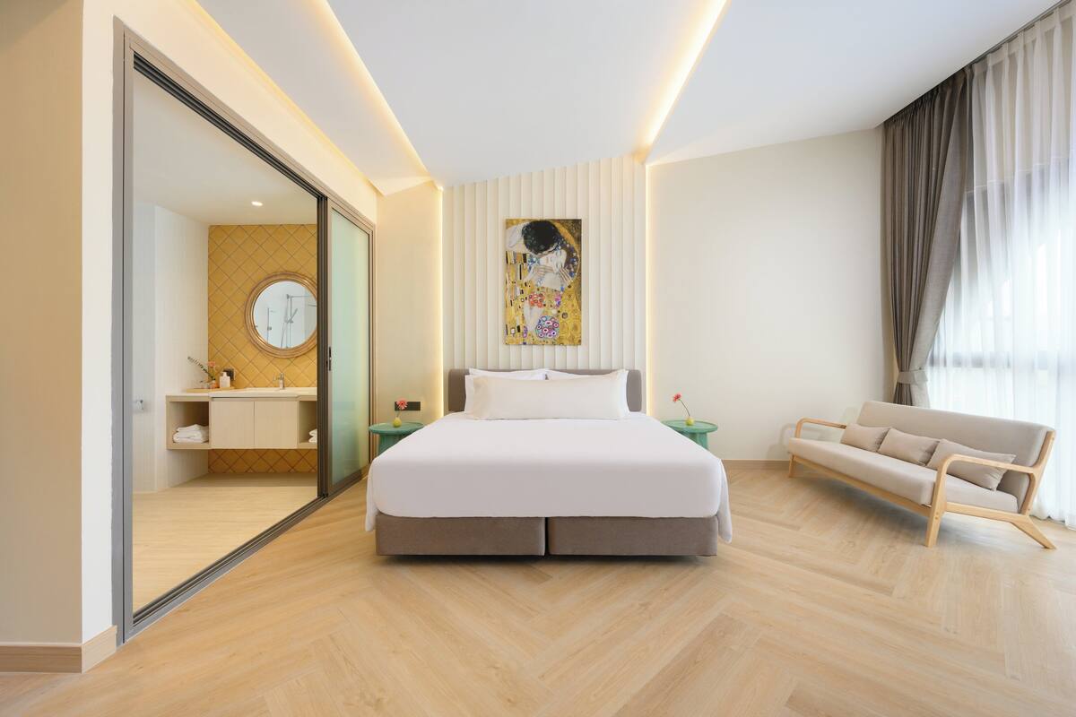 A modern hotel room with a large bed, a sofa, a bathroom with a mirror, and a painting on the wall.