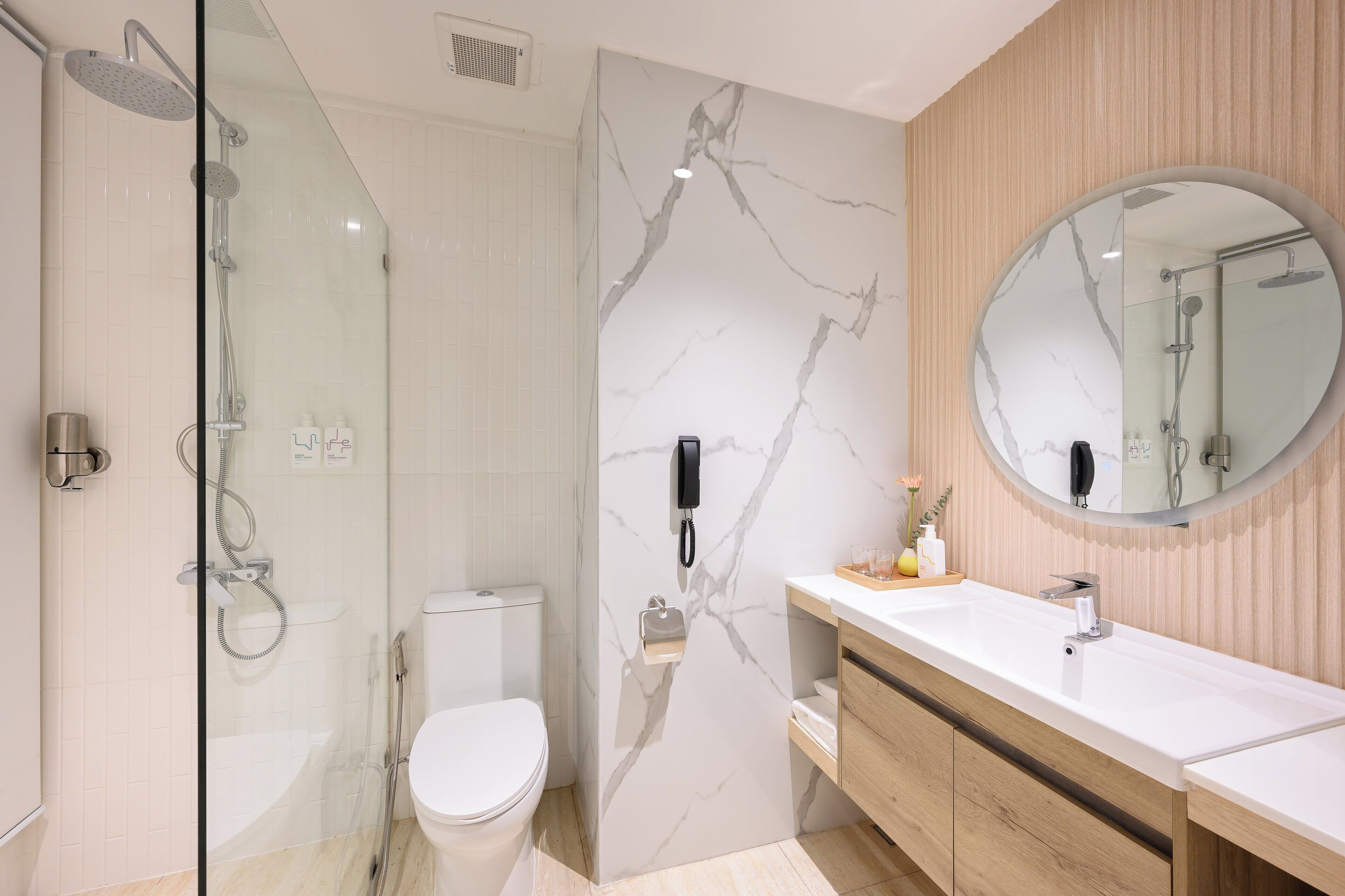 Un bagno moderno con cabina doccia in vetro, vaso bianco, parete in marmo, mobile lavabo in legno e specchio rotondo.