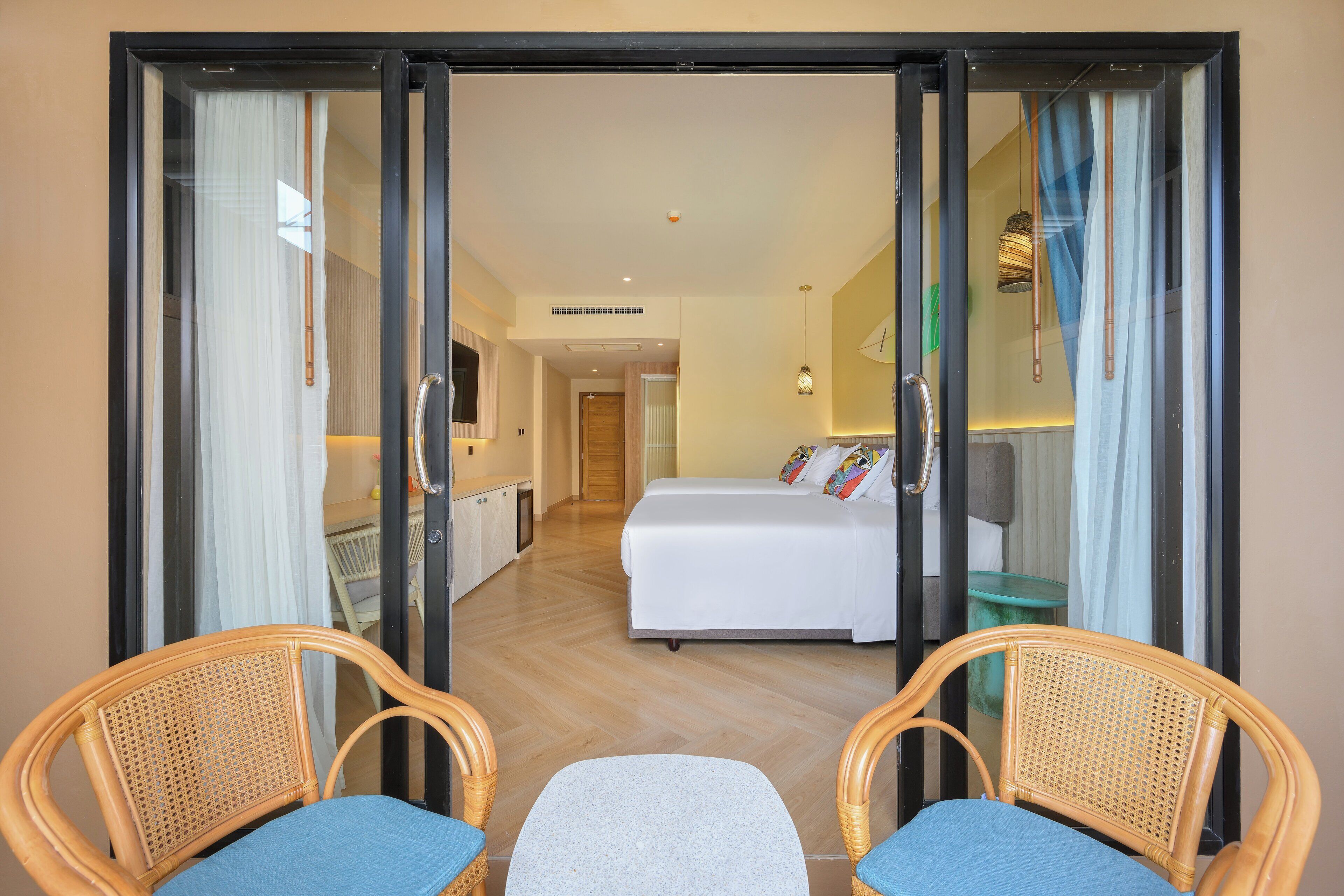Quarto de hotel com uma cama, duas cadeiras de vime, uma mesinha e uma porta de vidro deslizante para uma varanda.