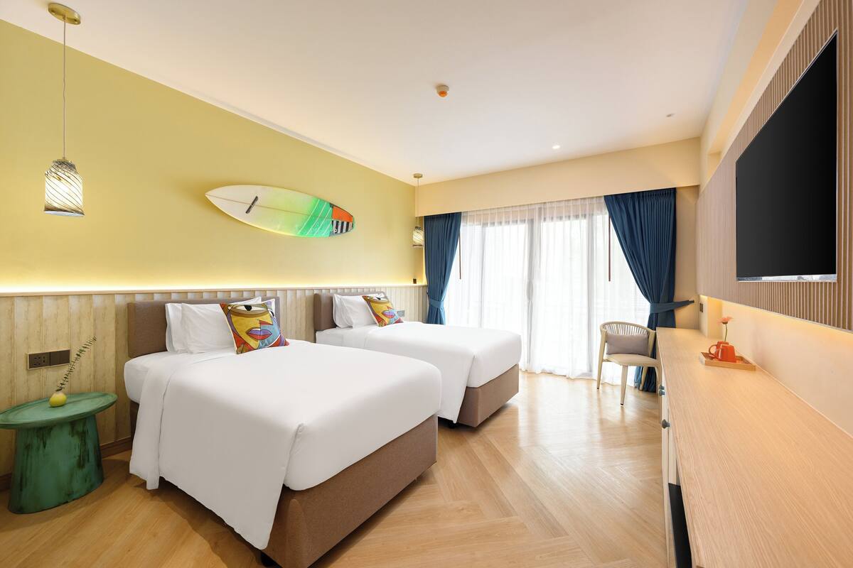 A hotel room with two beds, a desk, a TV, and a surfboard decoration on the wall.
