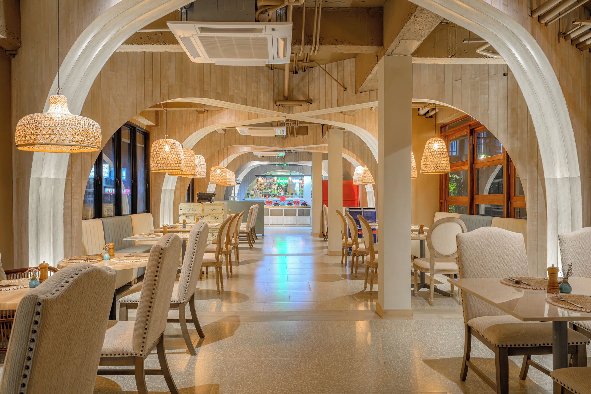 A spacious restaurant with high ceilings, arched wooden beams, and hanging woven light fixtures. Tables are arranged in the center, with chairs around them.