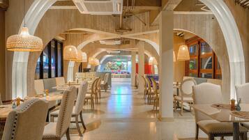 A spacious restaurant with high ceilings, arched wooden beams, and hanging woven light fixtures. Tables are arranged in the center, with chairs around them.
