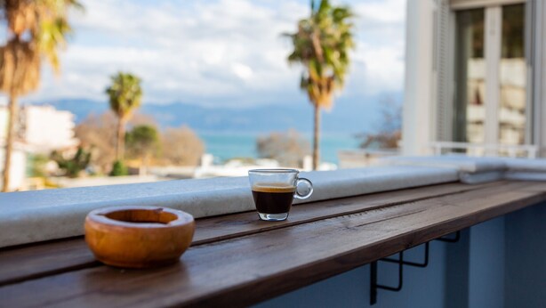 A wooden table with a glass of coffee and a wooden bowl on a balcony overlooking a coastal view with palm trees and mountains. - Lepanto Xenia Apartments 3F (Nafpaktos)