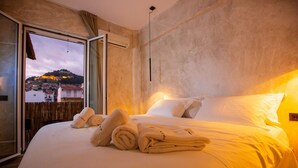 A hotel room with a bed, pillows, and towels. There is a window with a view of a cityscape and a mountain. - Lepanto Xenia Apartments 3F (Nafpaktos)