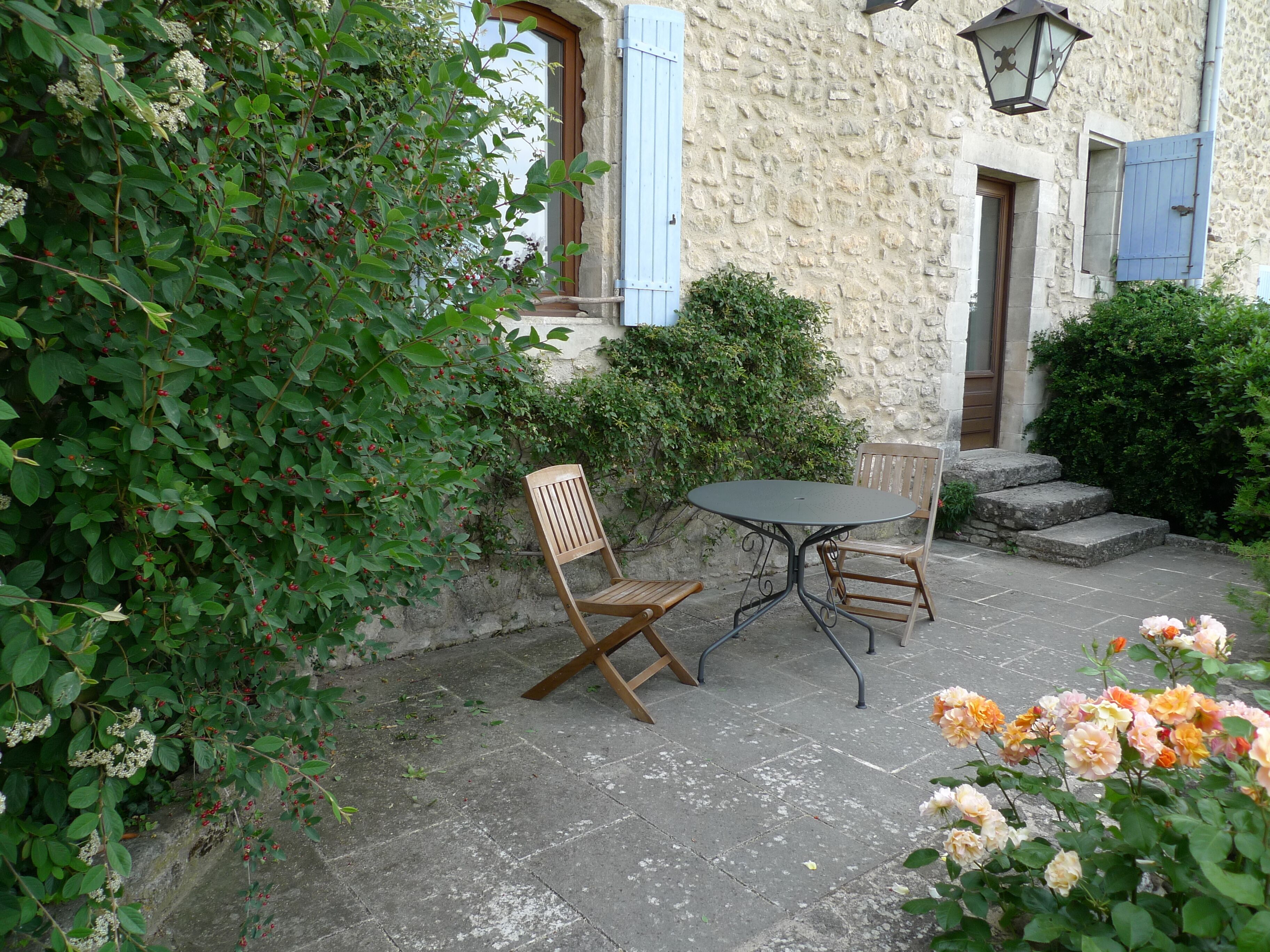 Une terrasse extérieure avec une table ronde et deux chaises en bois, entourée d’une végétation luxuriante et de plantes en fleurs.