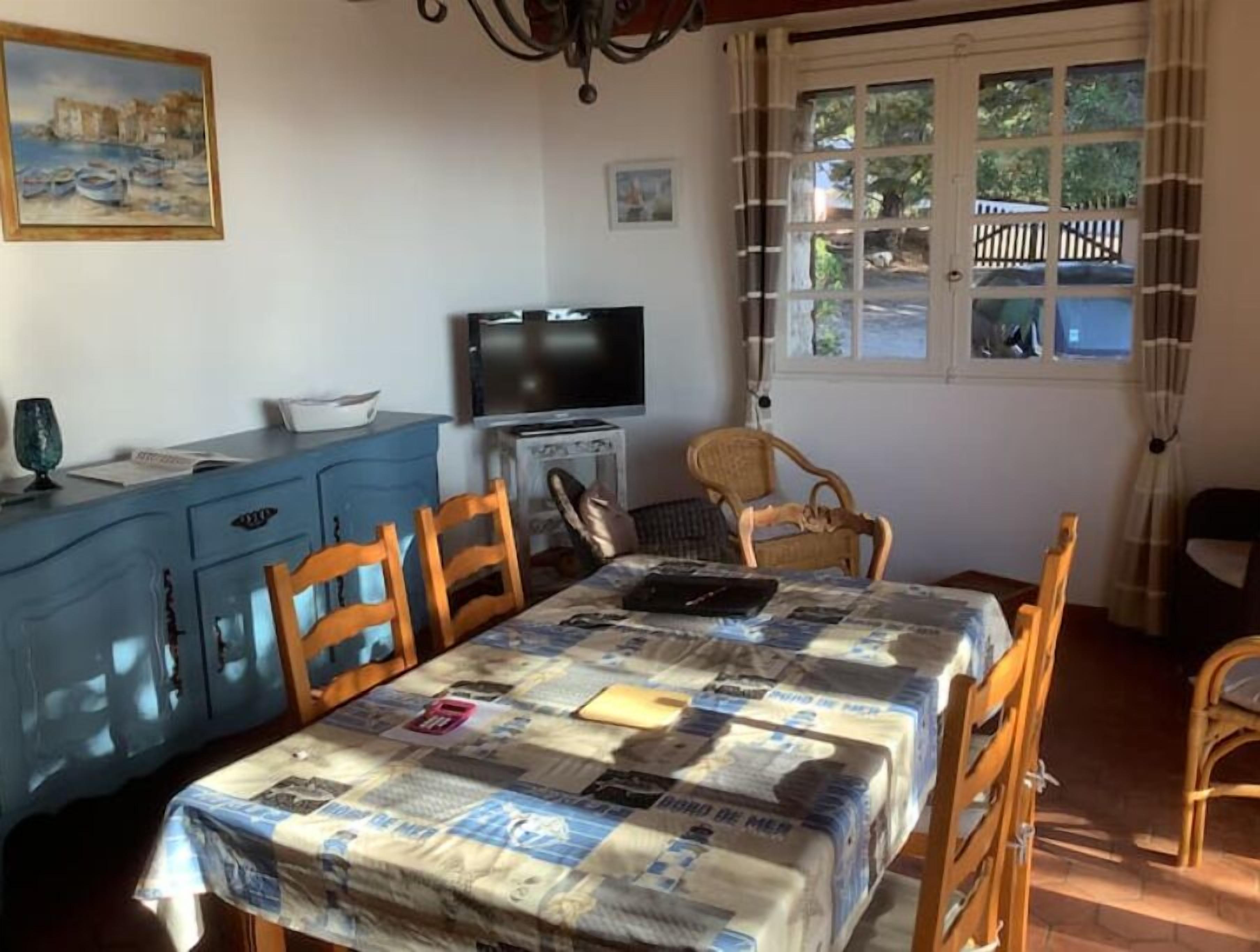 A dining area with a table covered by a patterned tablecloth, wooden chairs, a blue cabinet, a television, and a window with curtains.