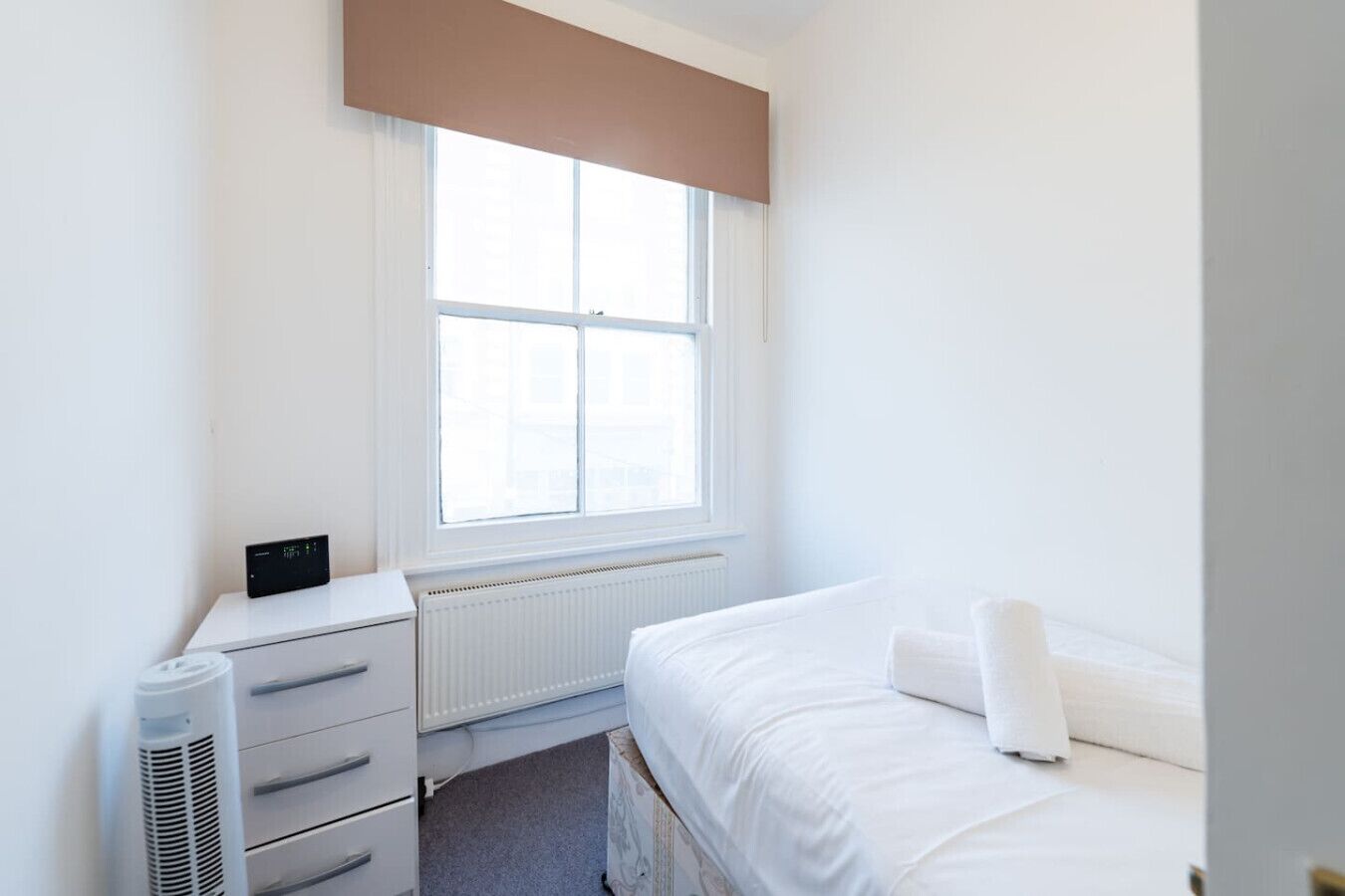 Un dormitorio con cama, radiador, calefactor, ventana con persianas y una mesita con un dispositivo.