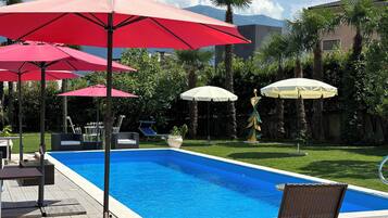A swimming pool with red and white umbrellas, lounge chairs, and a statue in a landscaped garden.