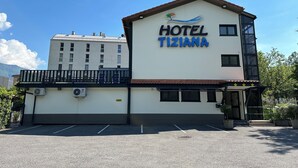 Un hotel llamado "HOTEL TIZIANA" con diseño moderno, que cuenta con un balcón y un estacionamiento frente a él.