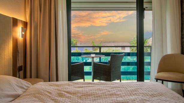 Habitación de hotel con cama, una mesita, dos sillas y un balcón con vistas a la puesta de sol.